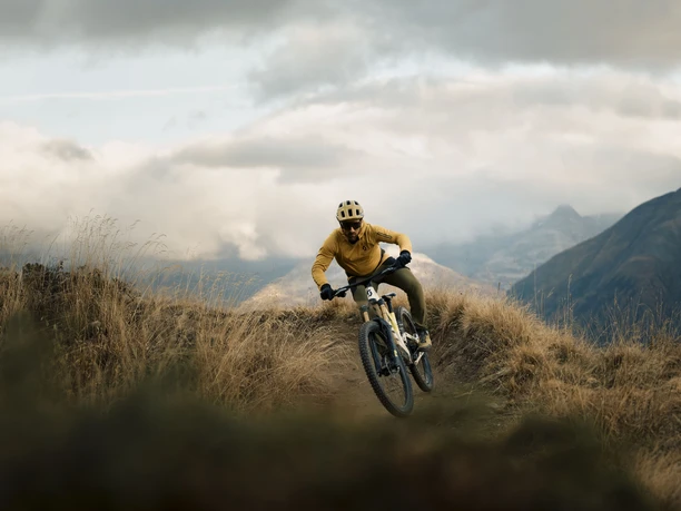 Dynamische Kurvenfahrt im alpinen Gelände Mountainbiker fährt auf dem Flowtrail Pass Wurzenbord dynamisch durch eine enge Kurve, umgeben von alpinen Wiesen und dramatischen Wolken.