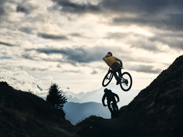 Temps de vol au-dessus du sentier alpin Un vététiste franchit un rebord de terrain sur le Flowtrail de Wurzenbord, tandis qu'un autre cycliste parcourt le parcours à travers l'Aletsch Arena.