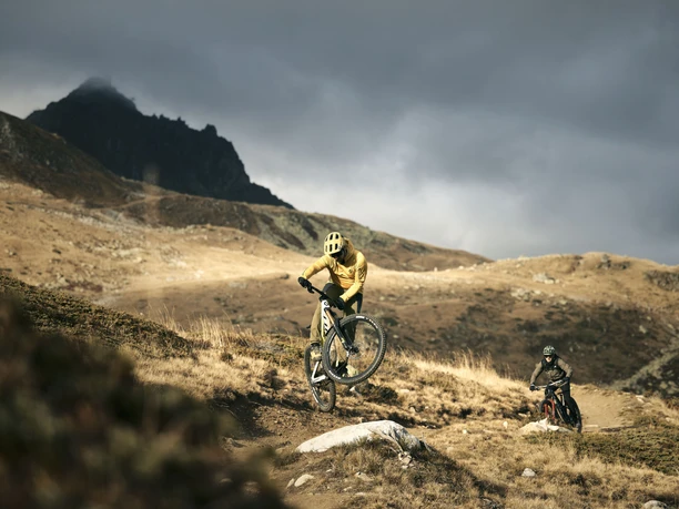 Action auf dem alpinen Flowtrail Mountainbiker fährt auf dem Flowtrail Wurzenbord einen Wheelie, während ein weiterer Fahrer auf dem alpinen Trail durch die Aletsch Arena folgt.