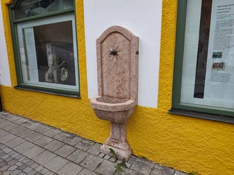 Brunnen-mit-Trinkwasser-Oberammergau-Museum.jpg Marmorbrunnen mit Trinkwasser an der gelb-weißen Fassade des Oberammergau Museums