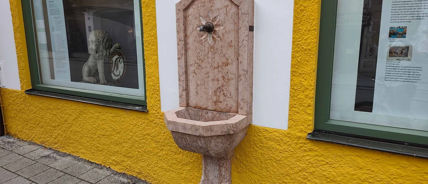 Brunnen-mit-Trinkwasser-Oberammergau-Museum.jpg Marmorbrunnen mit Trinkwasser an der gelb-weißen Fassade des Oberammergau Museums