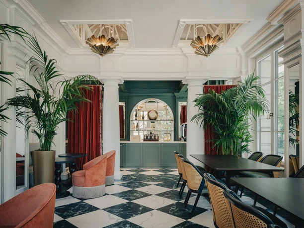 Taj Hessischer Hof Frankfurt- Elegantes Hotel-Interieur Elegantes Hotel-Interieur des Taj Hessischer Hof in Frankfurt mit schwarz-weißem Schachbrettboden, grüner Bar im Hintergrund, roten Samtvorhängen, Palmen und stilvollen Sitzbereichen.