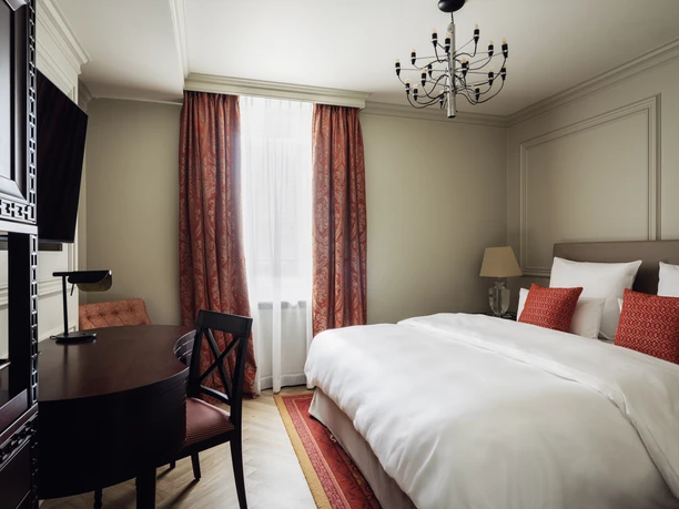 Taj Hessischer Hof Frankfurt- Elegantes Hotelzimmer im Taj Hessischer Hof Frankfurt Elegantes Hotelzimmer im Taj Hessischer Hof Frankfurt mit großem Doppelbett, roten Dekokissen und Vorhängen, einem dunklen Holzschreibtisch und einem modernen Kronleuchter.