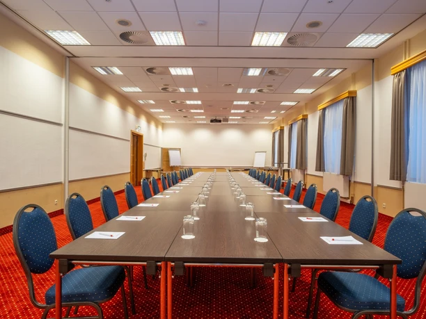 Airways Hotel Frankfurt Airport West Raunheim- Konferenzraum Konferenzraum im Airways Hotel Frankfurt Airport West mit einer langen, U-förmigen Tischreihe aus dunklem Holz, blauen Polsterstühlen, einem auffälligen roten Teppichboden und heller Deckenbeleuchtung.