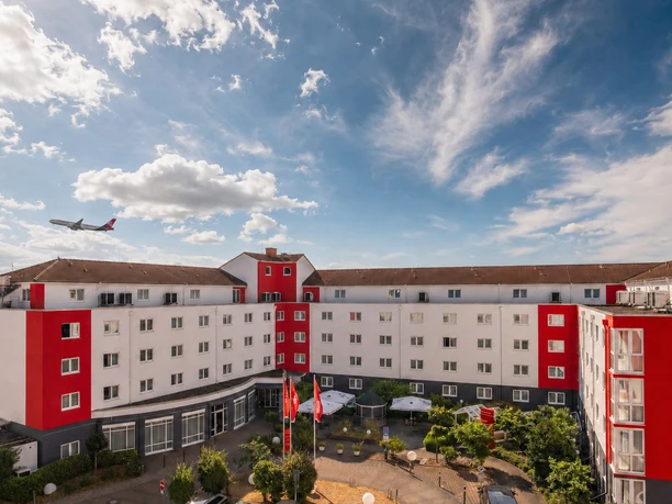 Airways Hotel Frankfurt Airport West Raunheim -Außenansicht Außenansicht des Airways Hotel Frankfurt Airport West in Raunheim mit markanter rot-weißer Fassade, einem gestalteten Innenhof mit Terrasse und einem fliegenden Flugzeug am blauen Himmel.
