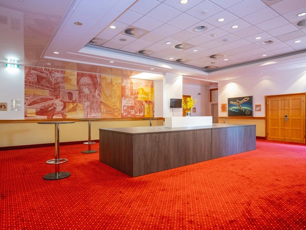 Airways Hotel Frankfurt Airport West Raunheim—Spacious Lobby Spacious lobby at the Airways Hotel Frankfurt Airport West, featuring bright red carpeting, a central wooden counter, bar tables, and a large mural with aviation motifs.