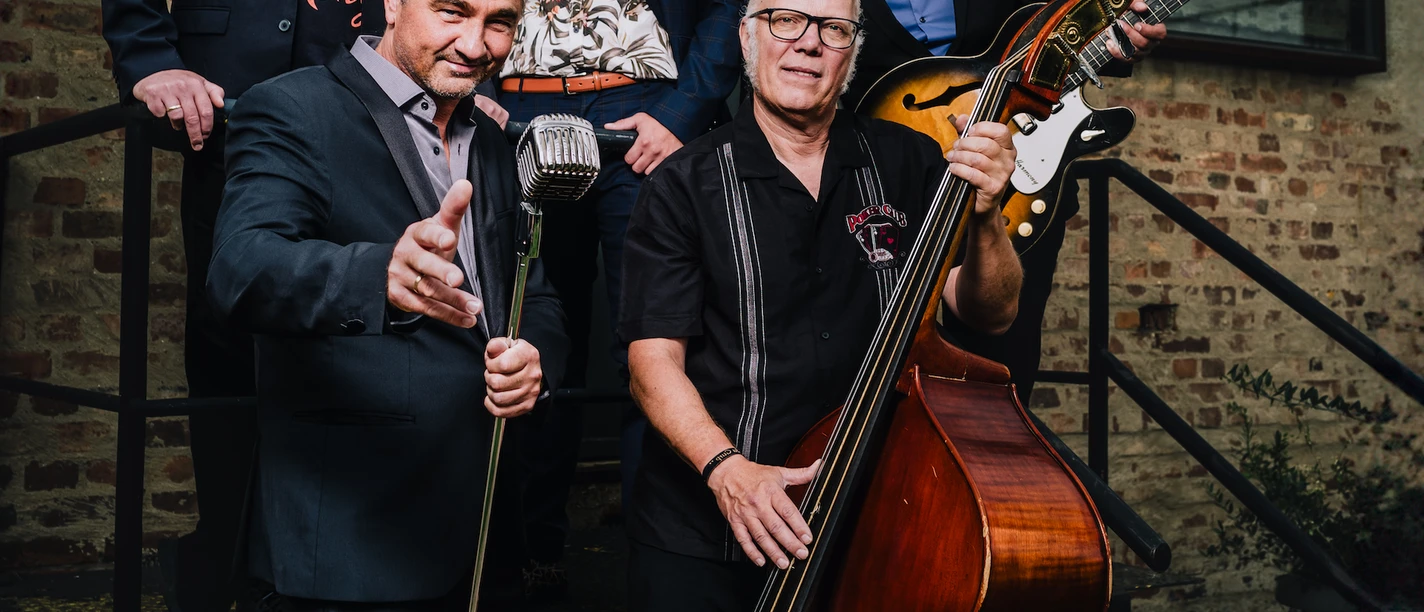 B.B. & The Blues Shacks posieren mit Mikrofon, Kontrabass und Gitarre vor Backsteinwand