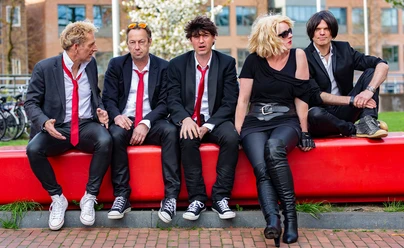 Band Back to Blondie sitzt in schwarzer Kleidung auf einer roten Bank im Freien.