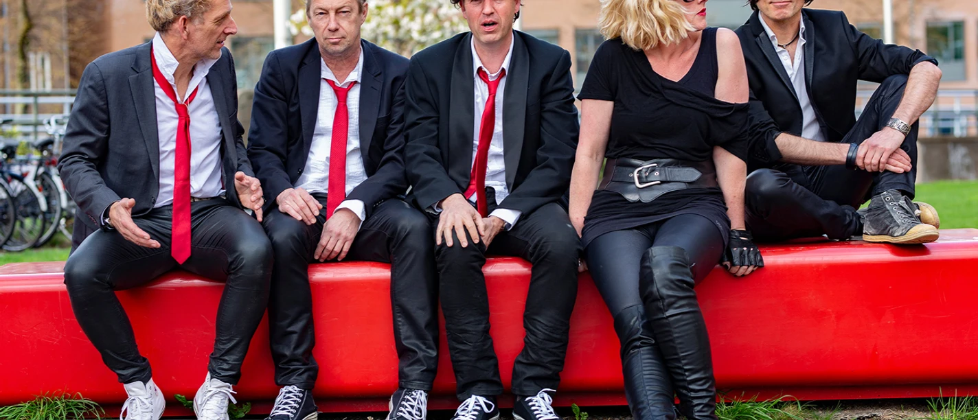 Back to Blondie.jpg Band Back to Blondie sitzt in schwarzer Kleidung auf einer roten Bank im Freien.