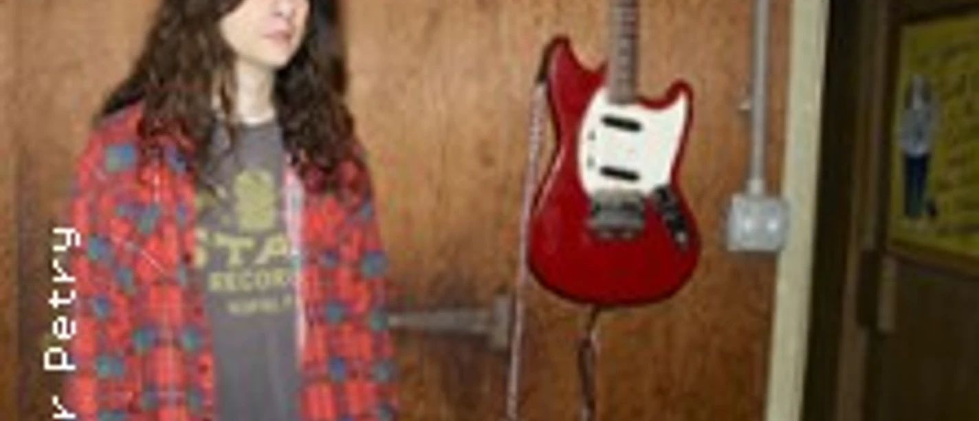 Kurt Vile steht vor einer Holzwand, daneben hängt eine rote E-Gitarre