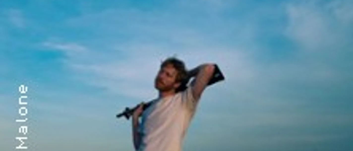 Max Fry mit Mikrofon am Ufer vor weitem blauem Himmel.