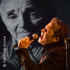 Sänger mit Mikrofon vor großem Schwarz-Weiß-Porträt von Charles Aznavour