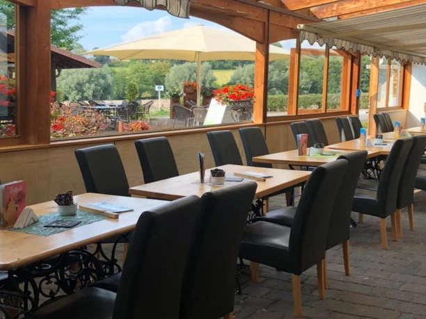 Sitzbereich des Cafés mit Polsterstühlen und Holztischen, im Hintergrund Fensterfront des Wintergartens und Blick auf den Garten