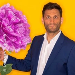 Christian Ehring im blauen Anzug vor gelbem Hintergrund mit großer pinker Blüte