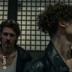 Zwei junge Männer in dunkler Szene, einer im Profil mit Lederjacke, einer im Hintergrund