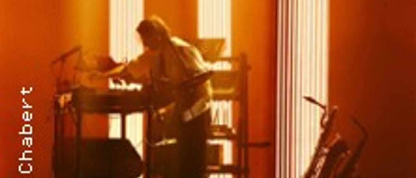 Musiker an Keyboard und Laptop auf orange beleuchteter Bühne, Gitarre am Rand