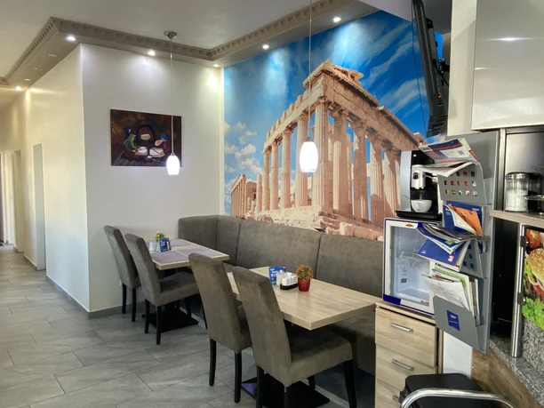 Athena_Grill_Innenansicht.jpg Innenraum Athena Grill mit grauen Sitzmöbeln und Wandbild eines antiken griechischen Tempels.