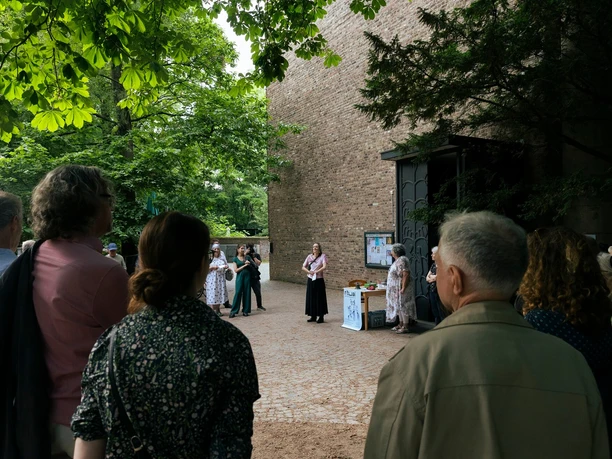ritual: Musik, Spaziergänge und Community Besuchergruppe vor Backsteinbau unter grünen Bäumen bei einem Musikspaziergang