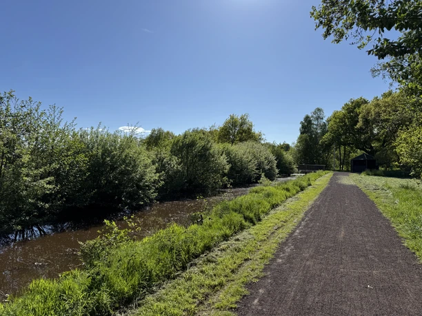Sommer am Landschaftspfad "Oll Wiek" Breiter Weg am Kanal mit Schutzhütte, Bäumen und Wiesen bei Sonnenschein.