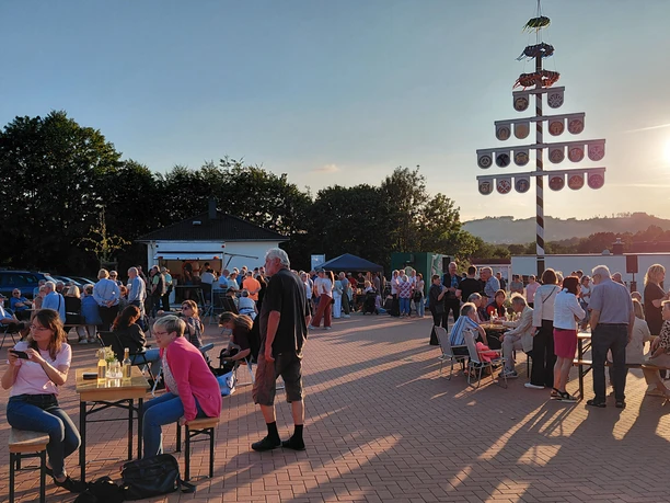 Menschen beim Beisammensein am geschmückten Maibaum auf dem Dorfplatz Hüllhorst im Abendlicht