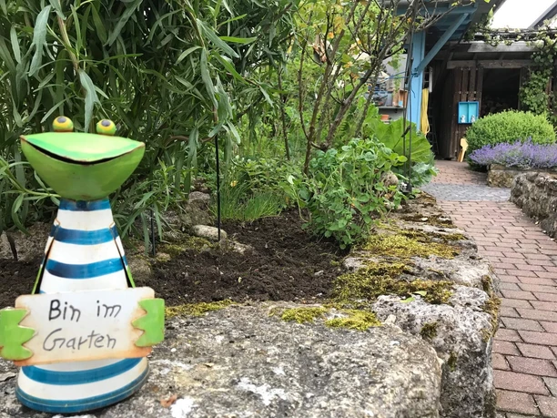 Willkommen im Gartenparadies Erhöhtes Kräuterbeet neben einem Weg, mit einer Metallgartendeko-Froschfigur, die ein Schild trägt, und einem Schuppen im Hintergrund