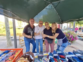 Vier Personen mit Sektgläsern an einem Flohmarktstand unter grünem Pavillon