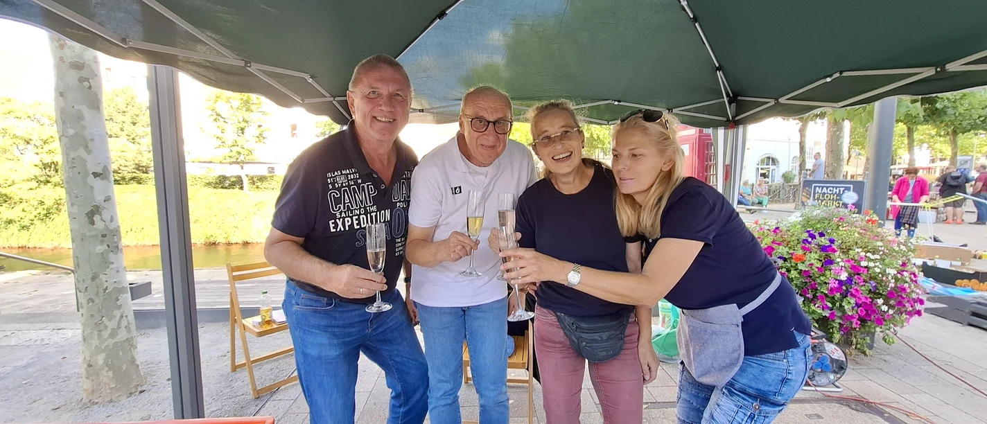 Vier Personen mit Sektgläsern an einem Flohmarktstand unter grünem Pavillon
