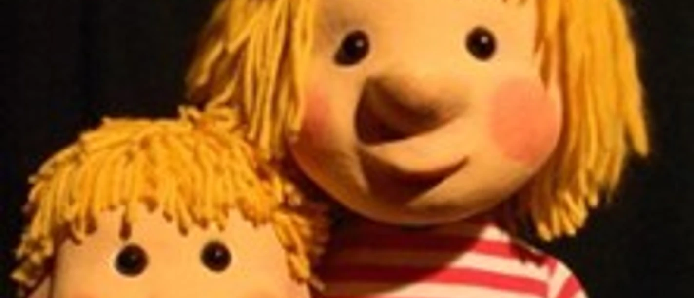 Zwei blonde Puppenfiguren, eine mit rot-weiß gestreiftem Shirt, vor schwarzem Hintergrund
