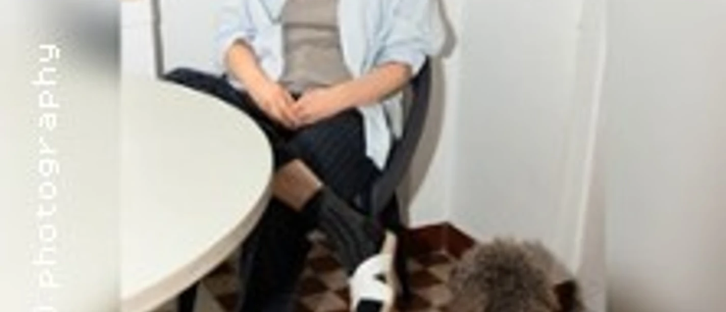 Eva Hegge sitzt auf einem Stuhl in hellem Hemd, neben ihr ein kleiner Hund.