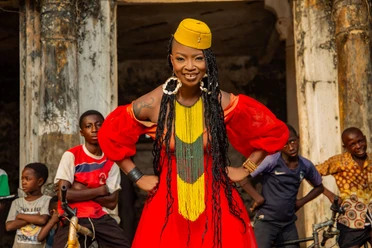 Dobet Gnahoré Dobet Gnahoré in rotem Kleid, umgeben von Kindern vor verwitterter Hausfassade