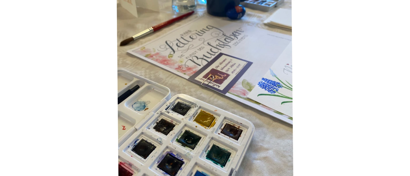 Handlettering-trifft-watercolor_bispingen.jpg Aquarellfarben, Pinsel und Handlettering-Vorlage auf einem Workshop-Tisch.