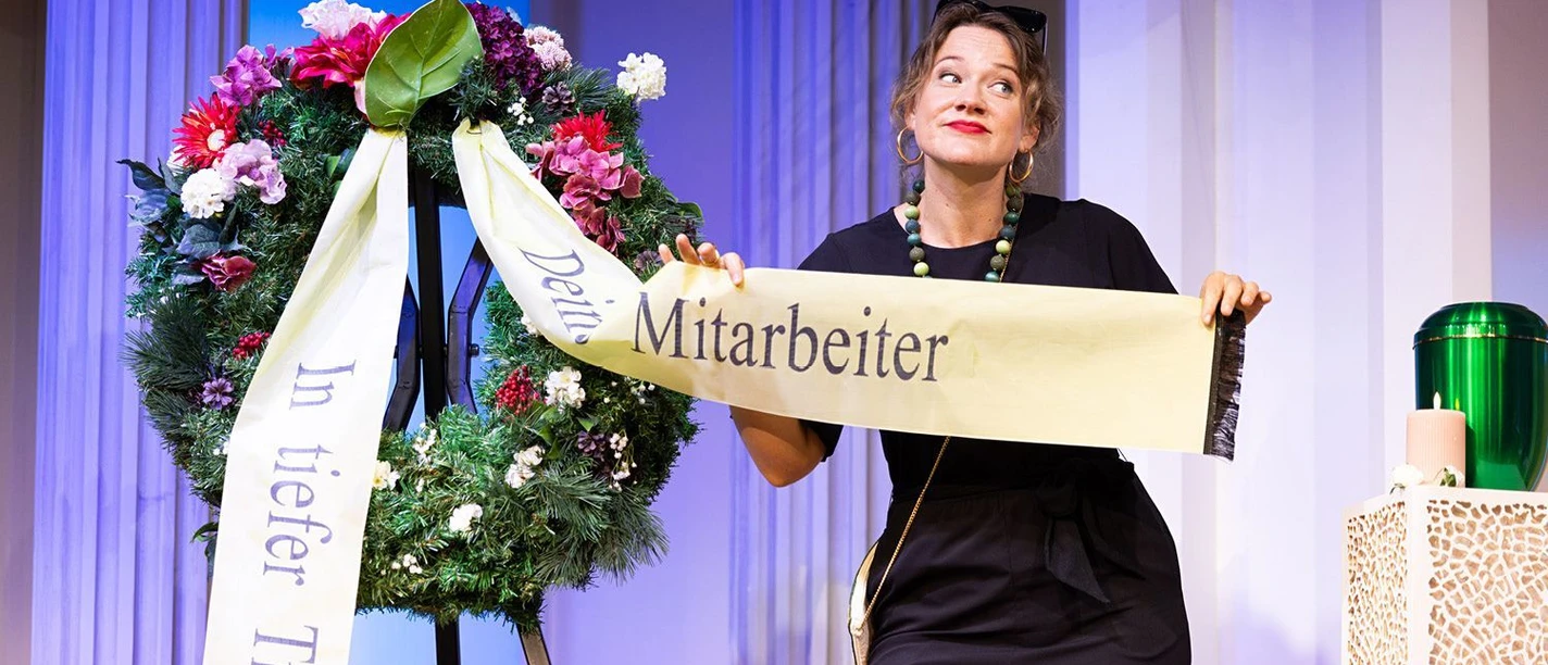Frau in Schwarz hält Trauerband mit Aufschrift Mitarbeiter neben einem Blumenkranz auf der Bühne.