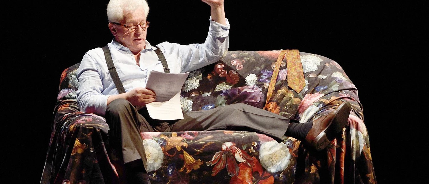 Schauspieler als Alexis Sorbas liest auf einem Blumensofa und hebt die Hand.