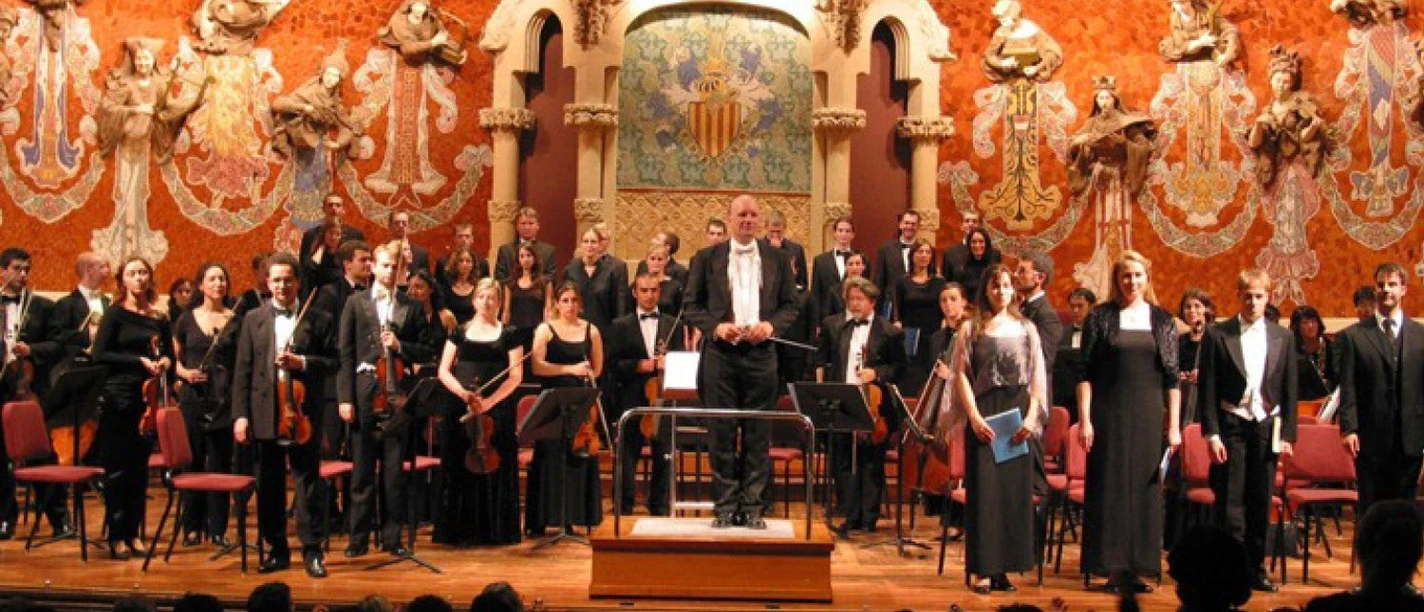 Orchester und Chor stehen nach einem Konzert gemeinsam mit dem Dirigenten auf einer festlich beleuchteten Bühne vor einem applaudierendem Publikum.