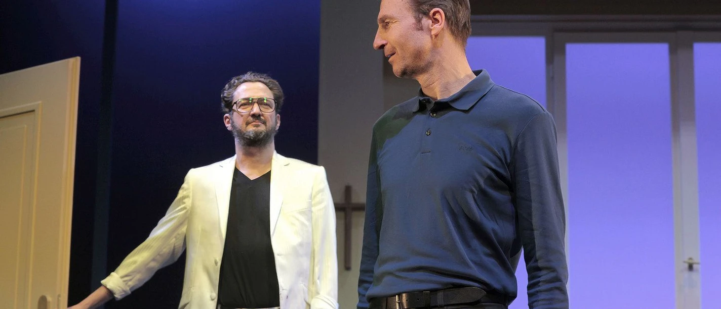 Szene aus dem Theaterstück „Einszweiundzwanzig vor dem Ende“: Zwei Schauspieler stehen sich auf einer Bühne mit Tür, Kreuz und blau beleuchtetem Bühnenbild gegenüber.