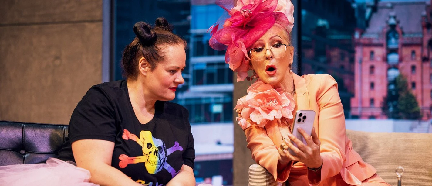 Zwei Schauspielerinnen sitzen auf einer Theaterbühne nebeneinander auf einem Sofa. Eine Frau in auffälligem rosa Kostüm und großem Hut hält ein Smartphone, während die andere interessiert auf den Bildschirm blickt.
