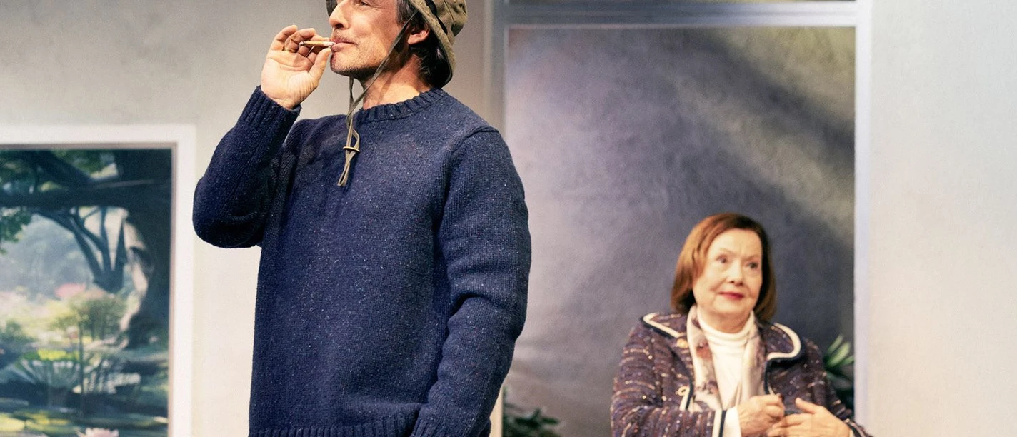 Mann mit Fischerhut und blauem Pullover raucht, daneben eine Frau in einer Theaterszene.