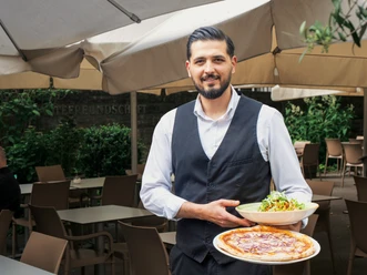 Restaurant Avanti 1995 Ein junger Kellner präsentiert zwei Gerichte auf Tellern. Auf dme einen ist ein Nudelgericht sichtbar, auf dem anderen ist eine Pizza abgebildet. Im Hintergrund ist eine Terrasse mit Stühlen und Tischen erkennbar.