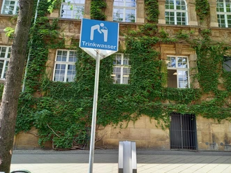 Sie sehen einen Schild mit der Aufschrift "Trinkbrunnen". Auf blauem Hintergrund ist eine Person abgebildet, die Wasser trinkt. Unterhalb des Schildes steht der Trinkbrunnen. Im Hintergrund ist die Fassade des Rathauses sichtbar.