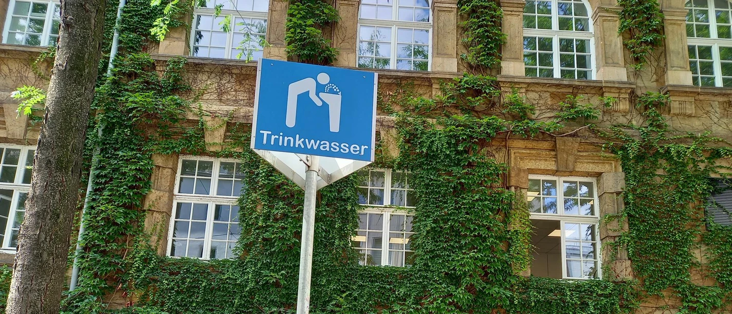 Trinkbrunnen Rathaus Sie sehen einen Schild mit der Aufschrift "Trinkbrunnen". Auf blauem Hintergrund ist eine Person abgebildet, die Wasser trinkt. Unterhalb des Schildes steht der Trinkbrunnen. Im Hintergrund ist die Fassade des Rathauses sichtbar.
