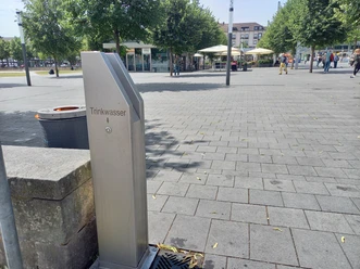 Trinkbrunnen Friedrichsplatz Der silberne Trinkbrunnen ist sichtbar. Über dem Knopf, befindet sich die Aufschrift "Trinkwasser" mit einem Pfeil zum Knopf. Im Hintergrund ist der Friedrichsplatz sichtbar mit dem Lokal Alex.