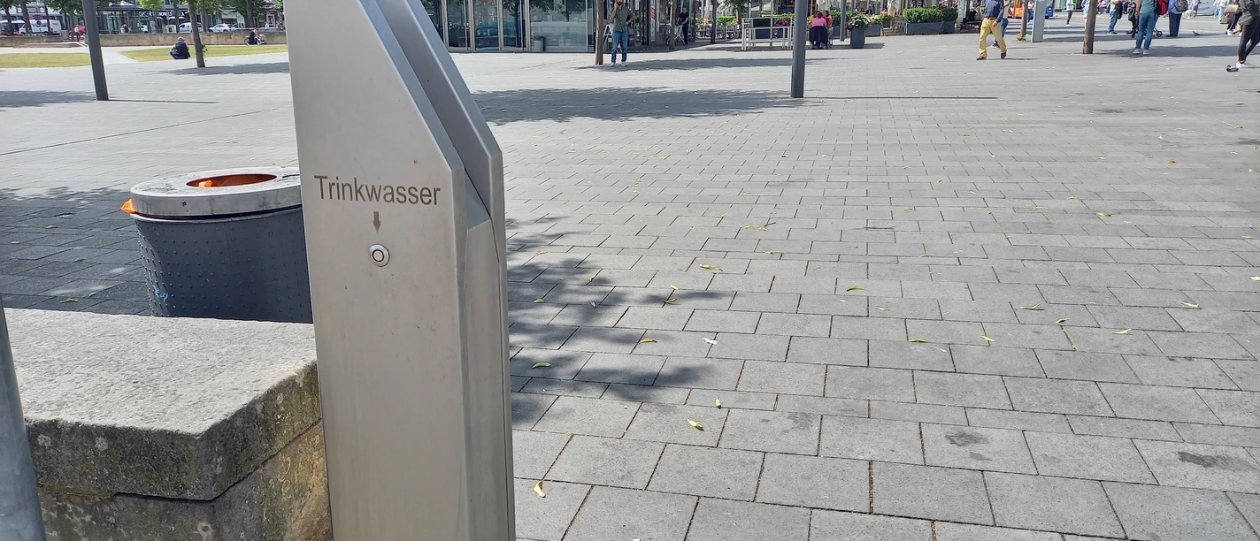 Trinkbrunnen Friedrichsplatz Der silberne Trinkbrunnen ist sichtbar. Über dem Knopf, befindet sich die Aufschrift "Trinkwasser" mit einem Pfeil zum Knopf. Im Hintergrund ist der Friedrichsplatz sichtbar mit dem Lokal Alex.