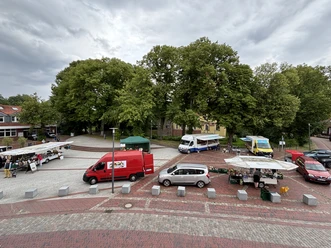 Wochenmarkt Pewsum Wochenmarkt in Pewsum mit Ständen, Fahrzeugen und großen Bäumen unter wolkigem Himmel.