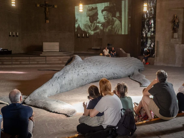 Eröffnung in St. Gertrud zur Ausstellung GESTRANDET Besucher sitzen vor einer großen Wal-Skulptur in der Ausstellung GESTRANDET in St. Gertrud