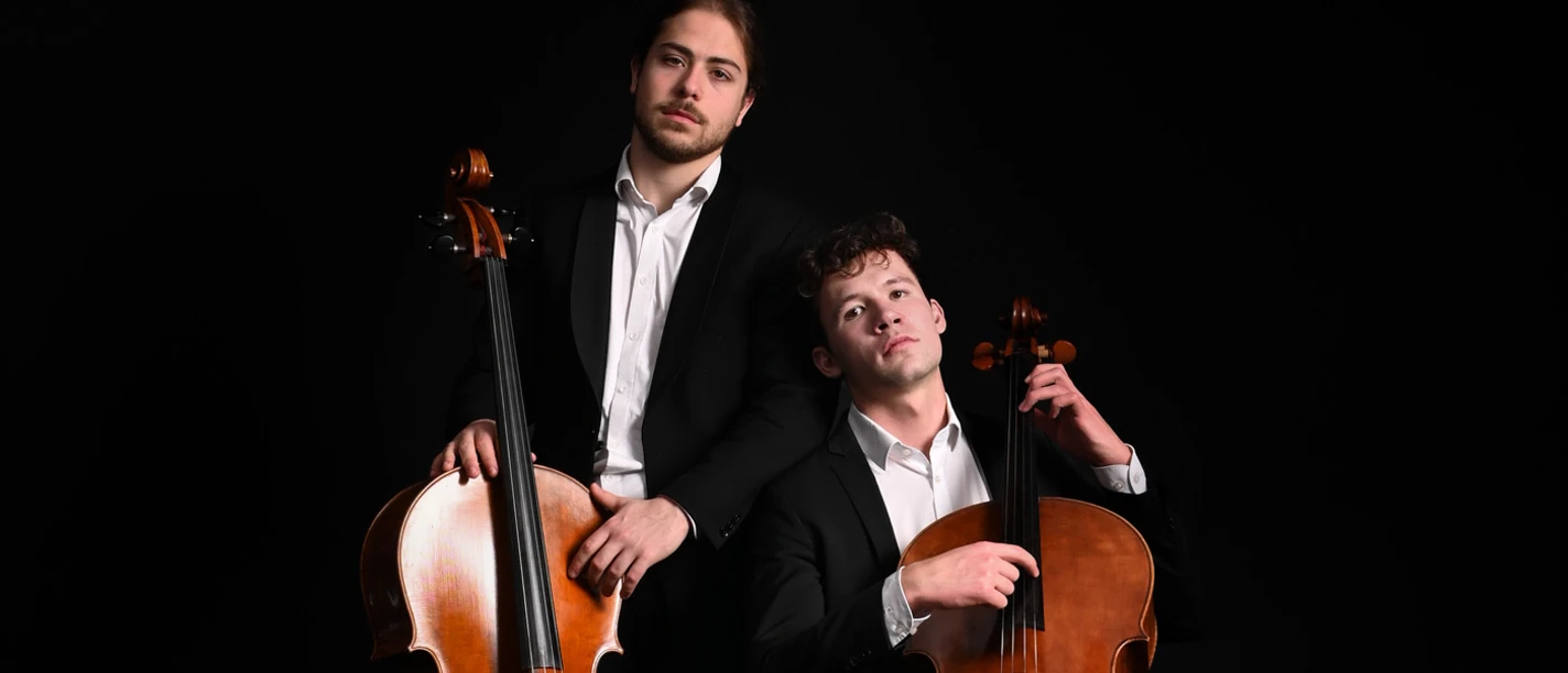 Zwei Musiker des CelloDuos in Anzügen posieren mit Celli vor schwarzem Hintergrund.