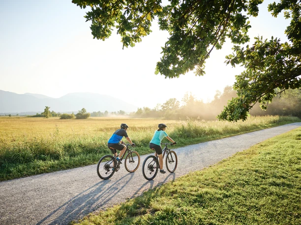 Zwei Radfahrende auf Kiesweg im Murnauer Moos, mit Wiesen, Bäumen und Bergblick