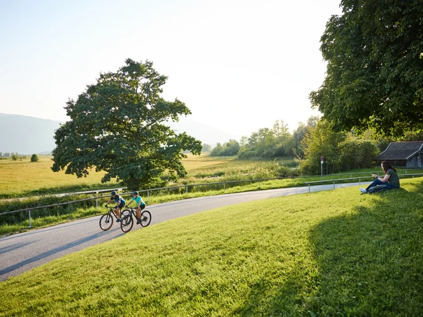 Zwei Radfahrer auf ruhiger Straße am Murnauer Moos, mit Wiesen, Bäumen und Bergen.