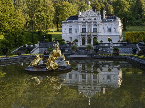 Schloss Linderhof mit goldenem Brunnen und Spiegelung im Wasser vor grünem Park