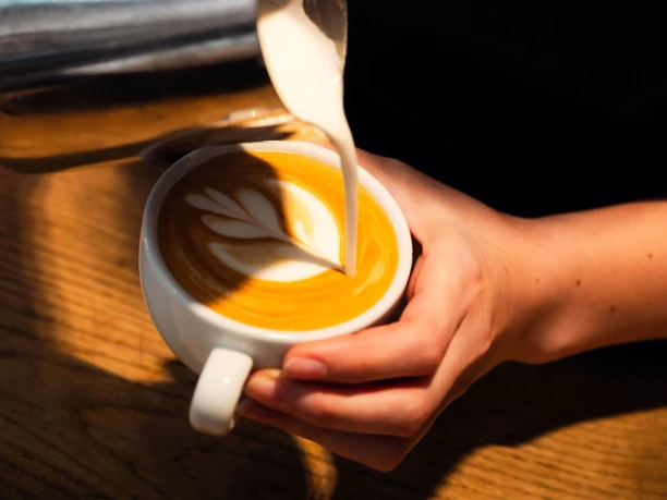 Barista gießt Milch in Cappuccino mit Latte-Art, moxxa-Logo auf der Schürze