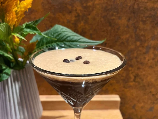 Espresso Martini im Martiniglas mit Crema, Kaffeebohnen und gelben Blüten im Hintergrund